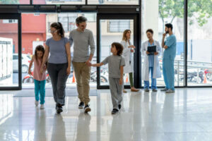 Happy family with two children walking into the hospital for an appointment with the pediatrician - Incidental workers at background