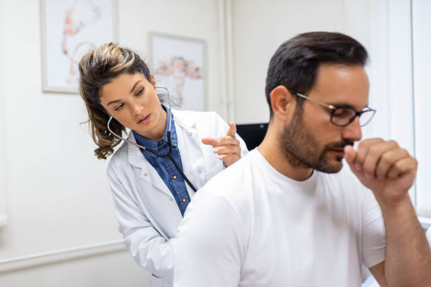 A female doctor at the clinic performs auscultation of the lungs of a patient with symptoms of coronavirus or pneumonia. He is coughing the doctor listens to the wheezing in the lungs