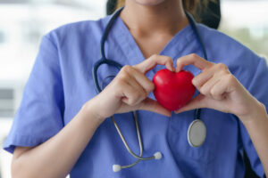 A nurse in blue scrubs holds a red heart in her hands, forming a heart shape around it, symbolizing heart disease prevention.
